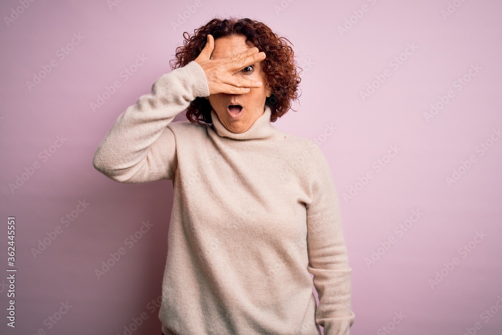 Middle age beautiful curly hair woman wearing casual turtleneck sweater over pink background peeking in shock covering face and eyes with hand, looking through fingers with embarrassed expression.