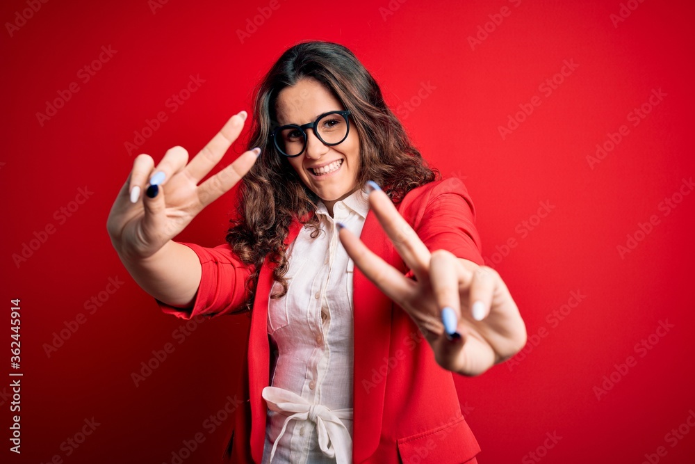 Young beautiful woman with curly hair wearing jacket and glasses over red background smiling looking to the camera showing fingers doing victory sign. Number two. Young beautiful woman with curly hair wearing jacket and glasses over red background smiling looking to the camera showing fingers doing victory sign. Number two.