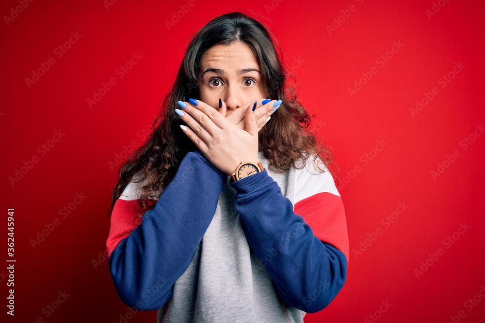 Young beautiful woman with curly hair wearing casual sweatshirt over isolated red background shocked covering mouth with hands for mistake. Secret concept. Young beautiful woman with curly hair wearing casual sweatshirt over isolated red background shocked covering mouth with hands for mistake. Secret concept.