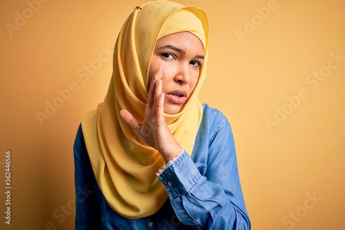 Young beautiful woman with curly hair wearing arab traditional hijab over yellow background hand on mouth telling secret rumor, whispering malicious talk conversation