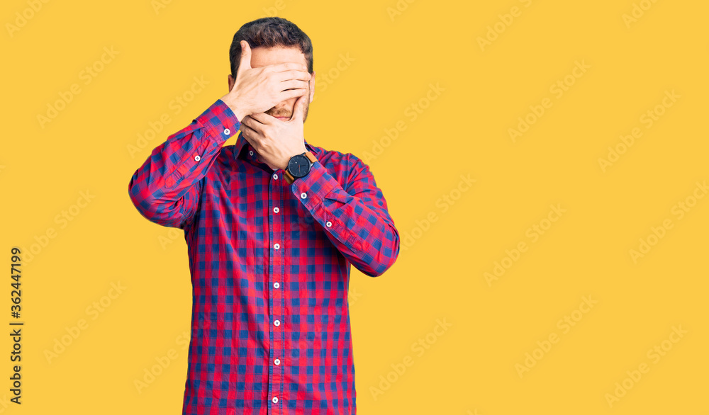 Handsome young man with bear wearing casual shirt covering eyes and mouth with hands, surprised and shocked. hiding emotion Handsome young man with bear wearing casual shirt covering eyes and mouth with hands, surprised and shocked. hiding emotion