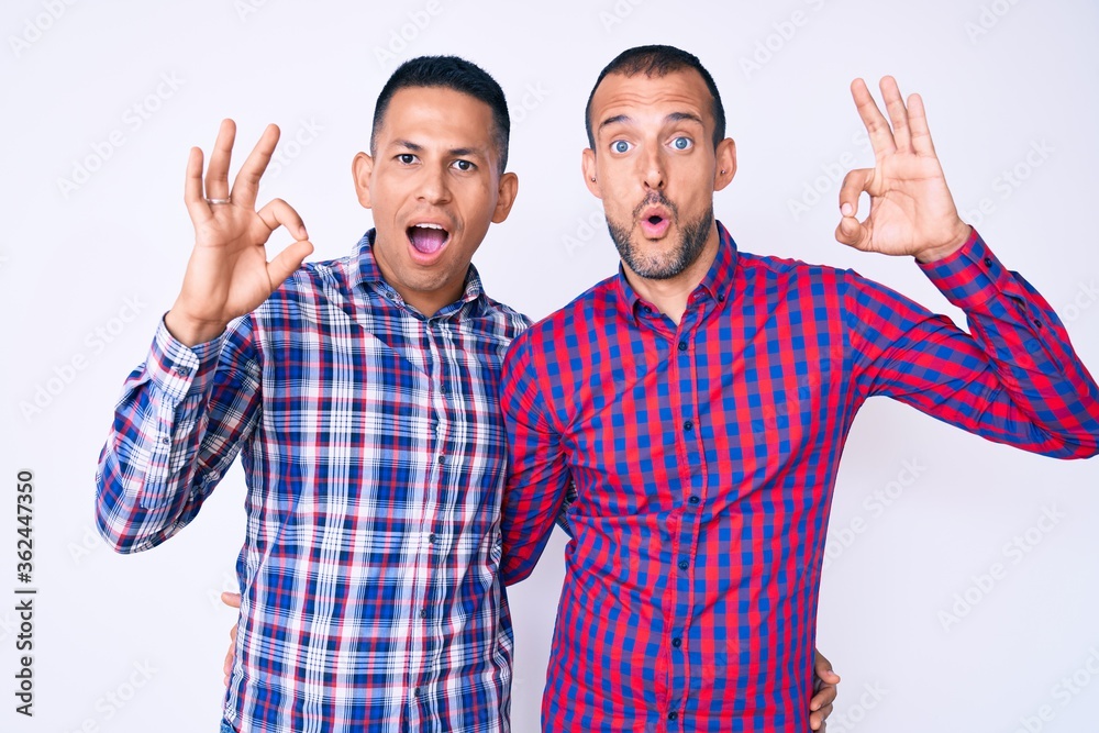 Naklejka premium Young gay couple of two men wearing casual clothes looking surprised and shocked doing ok approval symbol with fingers. crazy expression