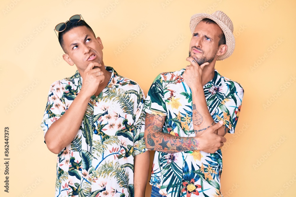 Young gay couple of two men wearing summer hat and hawaiian shirt thinking worried about a question, concerned and nervous with hand on chin Young gay couple of two men wearing summer hat and hawaiian shirt thinking worried about a question, concerned and nervous with hand on chin