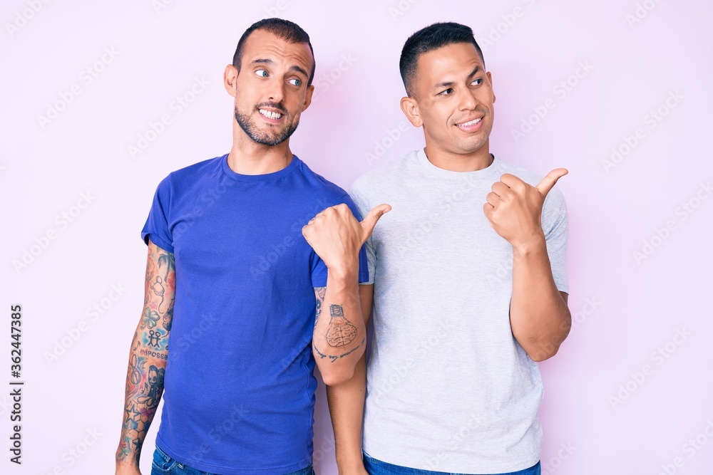 Young gay couple of two men wearing casual clothes smiling with happy face looking and pointing to the side with thumb up. Young gay couple of two men wearing casual clothes smiling with happy face looking and pointing to the side with thumb up.