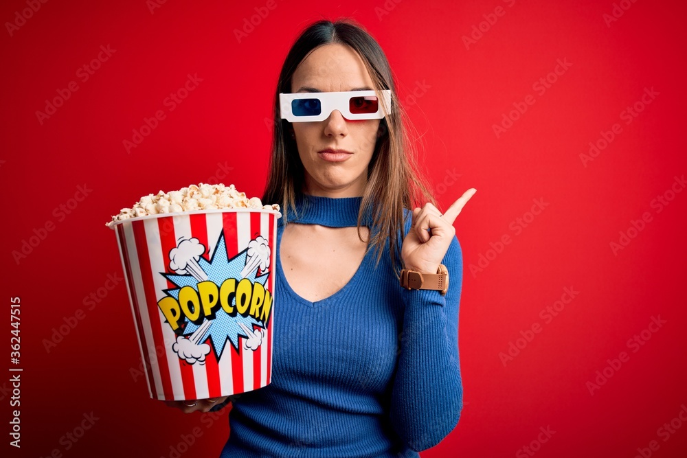 Obraz premium Young blonde woman wearing 3d glasses and eating pack of popcorn watching a movie on cinema Pointing with hand finger to the side showing advertisement, serious and calm face