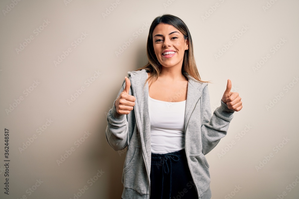 Young beautiful brunette sportswoman wearing sportswoman training over white background success sign doing positive gesture with hand, thumbs up smiling and happy. Cheerful expression. Young beautiful brunette sportswoman wearing sportswoman training over white background success sign doing positive gesture with hand, thumbs up smiling and happy. Cheerful expression.