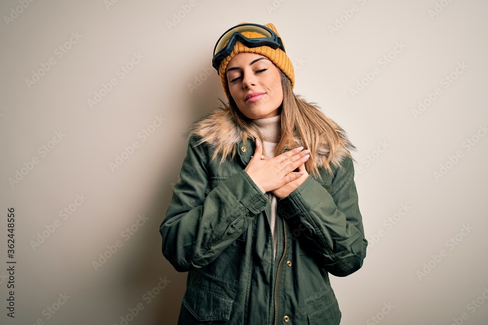 Young brunette skier woman wearing snow clothes and ski goggles over white background smiling with hands on chest with closed eyes and grateful gesture on face. Health concept.
