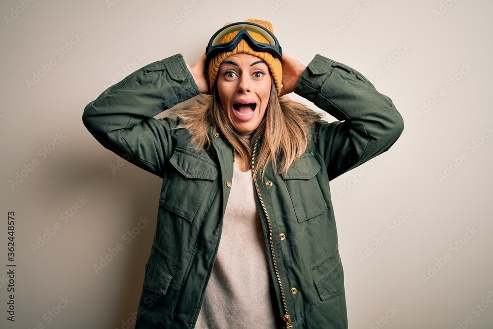 Young brunette skier woman wearing snow clothes and ski goggles over white background Crazy and scared with hands on head, afraid and surprised of shock with open mouth Young brunette skier woman wearing snow clothes and ski goggles over white background Crazy and scared with hands on head, afraid and surprised of shock with open mouth