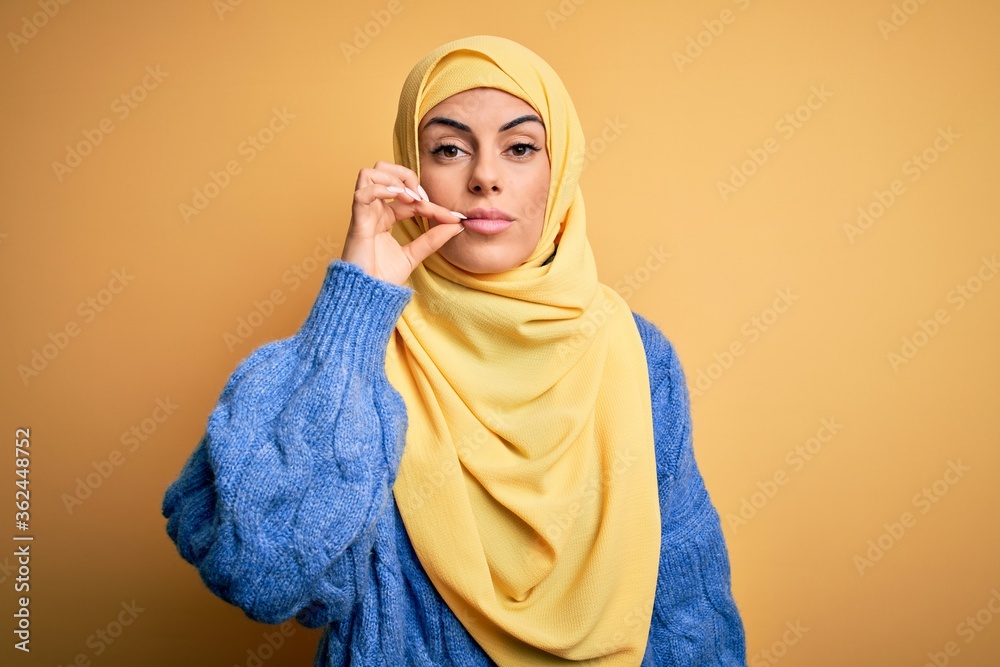Young beautiful brunette muslim woman wearing arab hijab over isolated yellow background mouth and lips shut as zip with fingers. Secret and silent, taboo talking Young beautiful brunette muslim woman wearing arab hijab over isolated yellow background mouth and lips shut as zip with fingers. Secret and silent, taboo talking