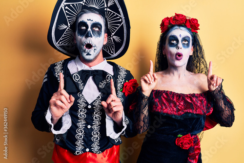 Young couple wearing mexican day of the dead costume over yellow amazed and surprised looking up and pointing with fingers and raised arms.