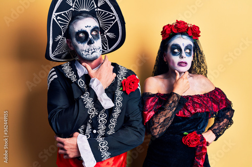 Young couple wearing mexican day of the dead costume over yellow thinking worried about a question, concerned and nervous with hand on chin