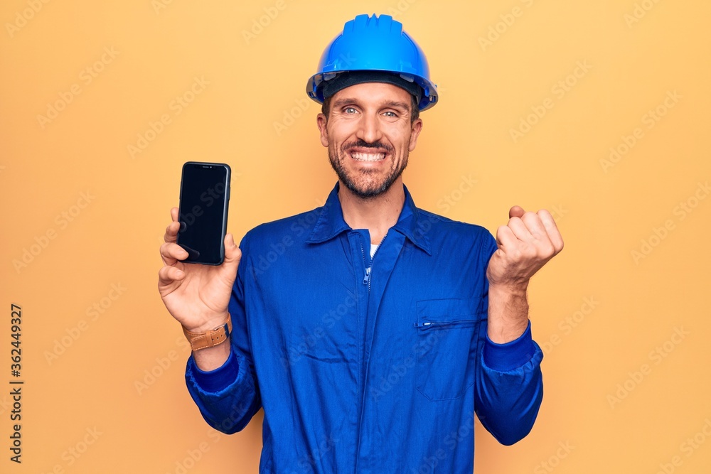 Young handsome worker man wearing uniform and hardhat holding smartphone showing screen screaming proud, celebrating victory and success very excited with raised arm Young handsome worker man wearing uniform and hardhat holding smartphone showing screen screaming proud, celebrating victory and success very excited with raised arm