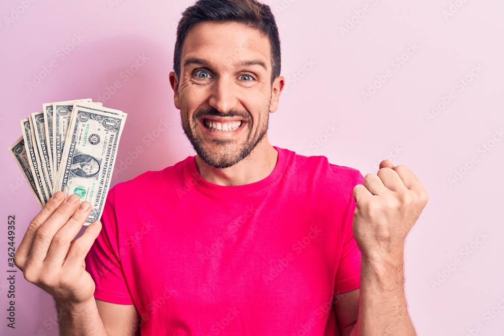 Young handsome man holding bunch of dollars banknotes over isolated pink background screaming proud, celebrating victory and success very excited with raised arm Young handsome man holding bunch of dollars banknotes over isolated pink background screaming proud, celebrating victory and success very excited with raised arm