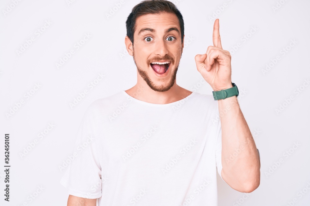 Young handsome man wearing casual white tshirt pointing finger up with successful idea. exited and happy. number one. Young handsome man wearing casual white tshirt pointing finger up with successful idea. exited and happy. number one.