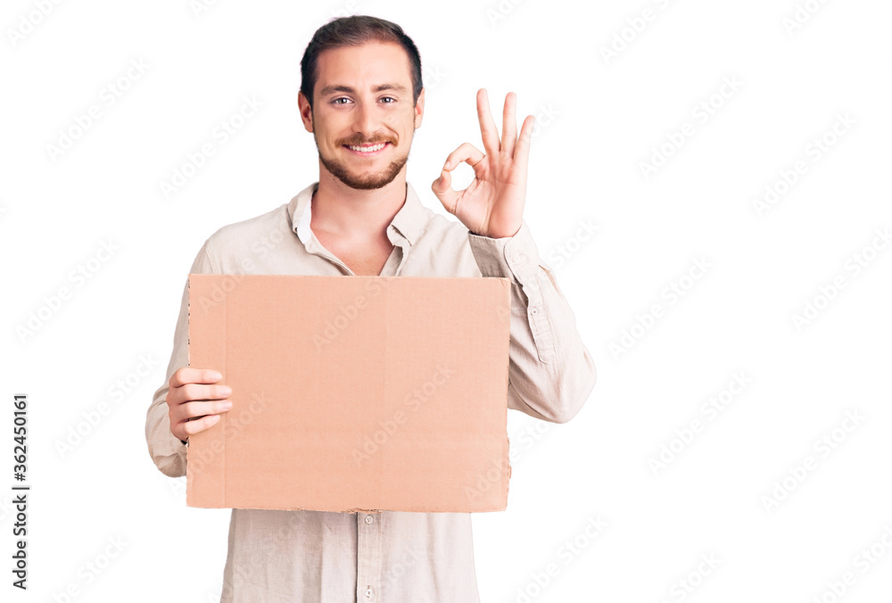 Young handsome caucasian man holding empty cardboard banner doing ok sign with fingers, smiling friendly gesturing excellent symbol Young handsome caucasian man holding empty cardboard banner doing ok sign with fingers, smiling friendly gesturing excellent symbol