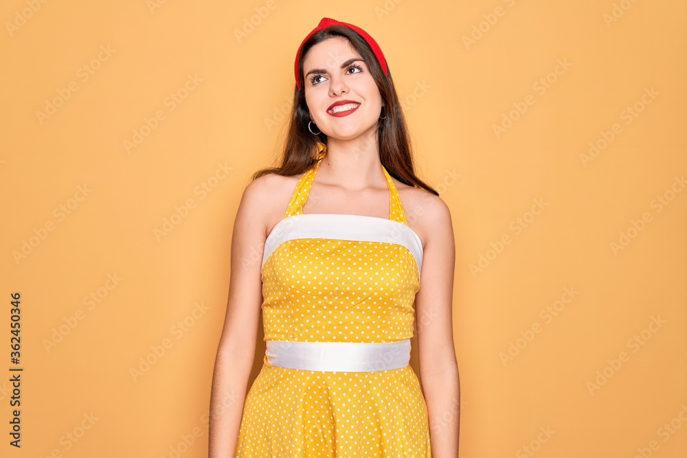 Φωτογραφία Young beautiful pin up woman wearing 50s fashion vintage dress over yellow background looking away to side with smile on face, natural expression
