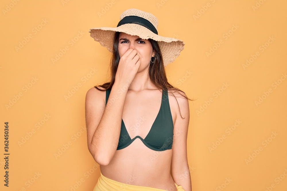 Young beautiful girl wearing swimwear bikini and summer sun hat over yellow background smelling something stinky and disgusting, intolerable smell, holding breath with fingers on nose. Bad smell Young beautiful girl wearing swimwear bikini and summer sun hat over yellow background smelling something stinky and disgusting, intolerable smell, holding breath with fingers on nose. Bad smell