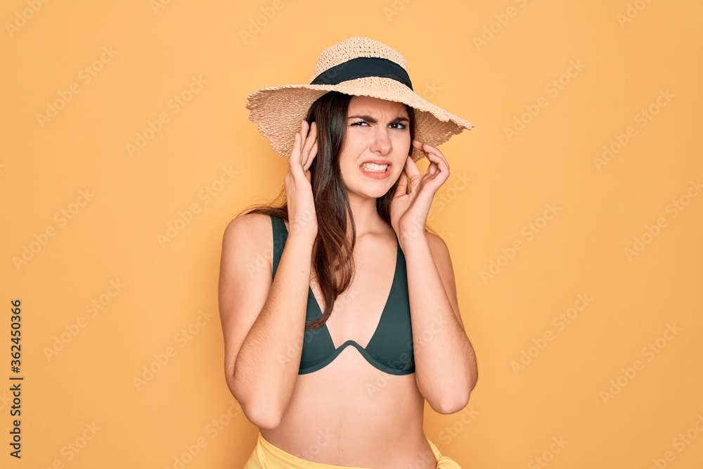 Φωτογραφία Young beautiful girl wearing swimwear bikini and summer sun hat over yellow background covering ears with fingers with annoyed expression for the noise of loud music