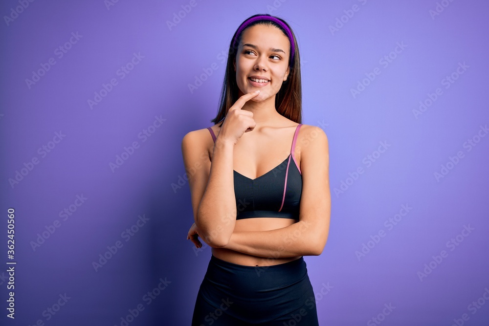 Young beautiful sporty girl doing sport wearing sportswear over isolated purple background looking confident at the camera smiling with crossed arms and hand raised on chin. Thinking positive. Young beautiful sporty girl doing sport wearing sportswear over isolated purple background looking confident at the camera smiling with crossed arms and hand raised on chin. Thinking positive.