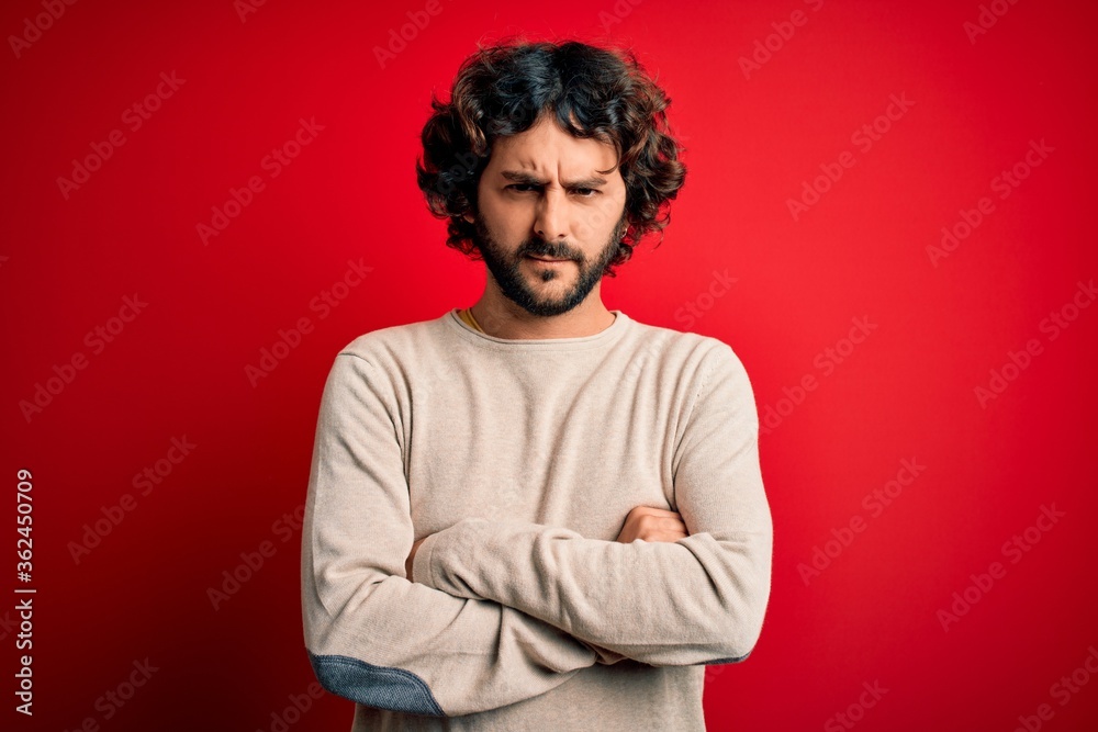 Young handsome man with beard wearing casual sweater standing over red background skeptic and nervous, disapproving expression on face with crossed arms. Negative person. Young handsome man with beard wearing casual sweater standing over red background skeptic and nervous, disapproving expression on face with crossed arms. Negative person.
