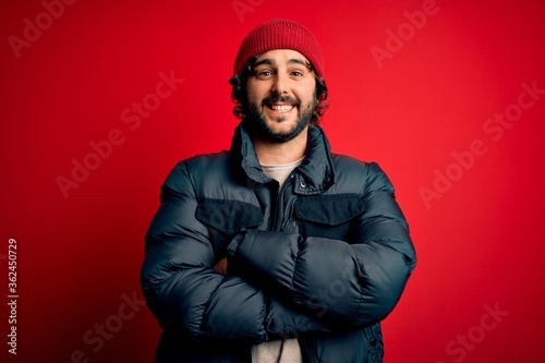 Young handsome man with beard wearing casual coat and wool cap over red background happy face smiling with crossed arms looking at the camera. Positive person. © Krakenimages.com