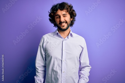 Young handsome business man with beard wearing shirt standing over purple background winking looking at the camera with sexy expression, cheerful and happy face. © Krakenimages.com