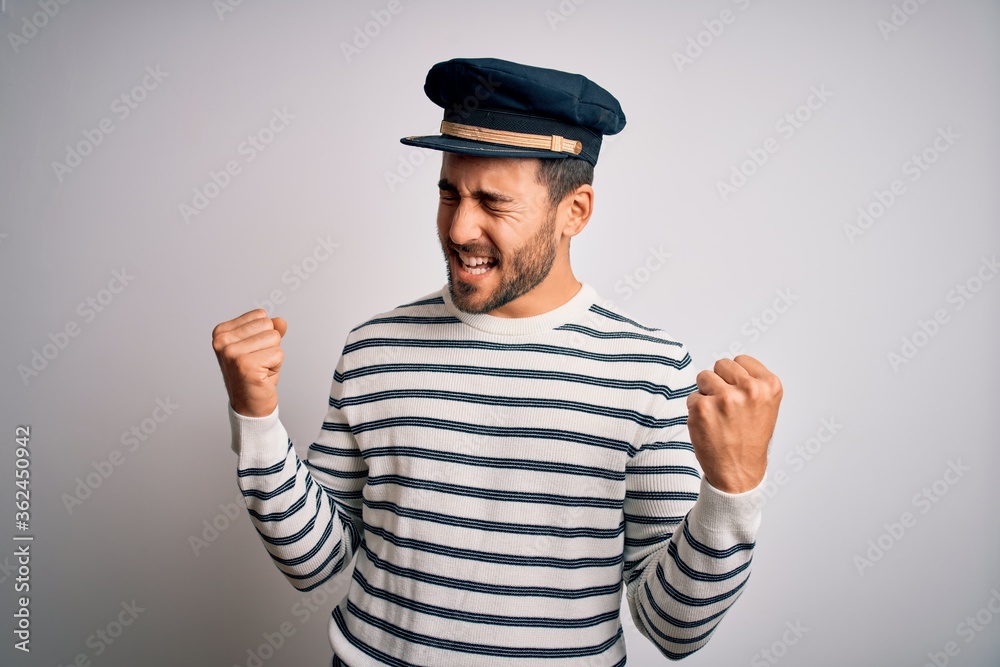 Young handsome sailor man with beard wearing navy striped uniform and captain hat very happy and excited doing winner gesture with arms raised, smiling and screaming for success. Celebration concept. Young handsome sailor man with beard wearing navy striped uniform and captain hat very happy and excited doing winner gesture with arms raised, smiling and screaming for success. Celebration concept.