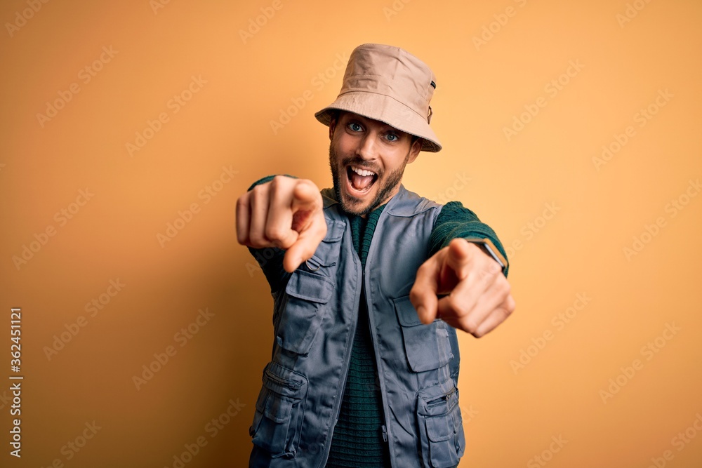 Handsome tourist man with beard on vacation wearing explorer hat over yellow background pointing to you and the camera with fingers, smiling positive and cheerful Handsome tourist man with beard on vacation wearing explorer hat over yellow background pointing to you and the camera with fingers, smiling positive and cheerful