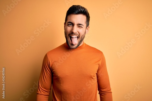 Young handsome man with beard wearing casual sweater standing over yellow background sticking tongue out happy with funny expression. Emotion concept.