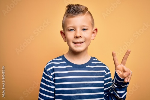 Young little caucasian kid with blue eyes wearing nautical striped shirt over yellow background smiling with happy face winking at the camera doing victory sign. Number two.