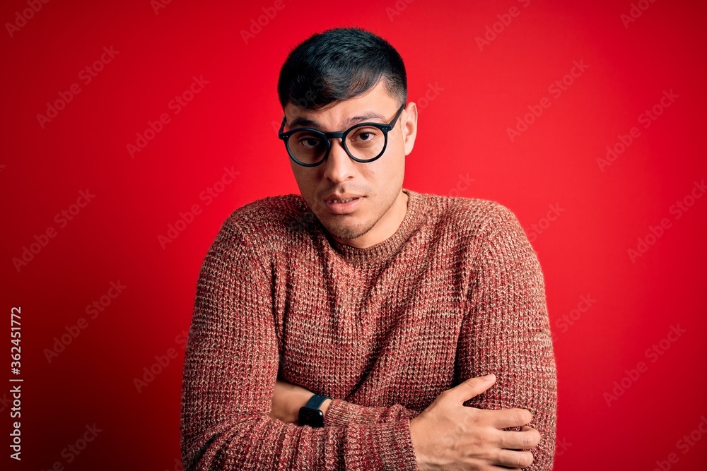 Young handsome hispanic man wearing nerd glasses over red background shaking and freezing for winter cold with sad and shock expression on face Young handsome hispanic man wearing nerd glasses over red background shaking and freezing for winter cold with sad and shock expression on face