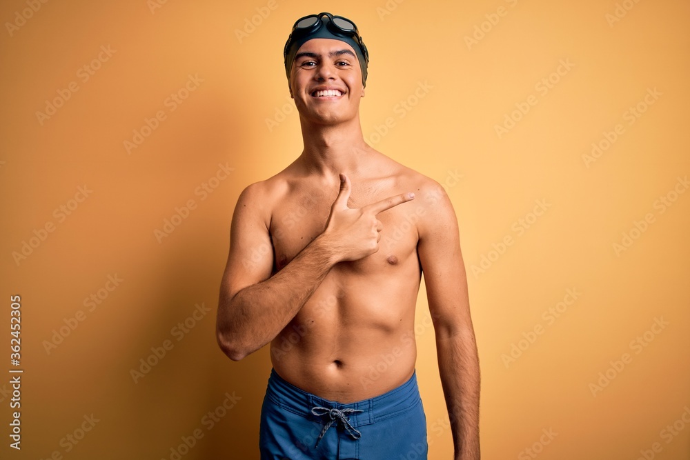 Young handsome man shirtless wearing swimsuit and swim cap over isolated yellow background cheerful with a smile on face pointing with hand and finger up to the side with happy and natural expression