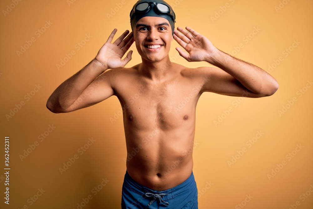 Young handsome man shirtless wearing swimsuit and swim cap over isolated yellow background Smiling pulling ears with fingers, funny gesture. Audition problem