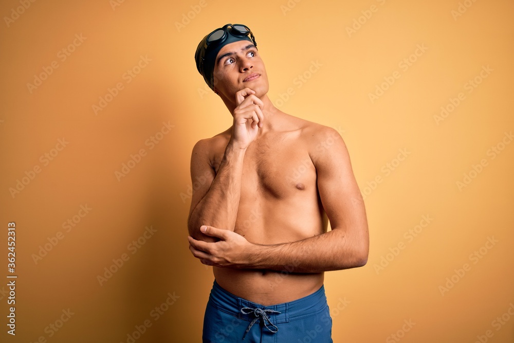Young handsome man shirtless wearing swimsuit and swim cap over isolated yellow background with hand on chin thinking about question, pensive expression. Smiling and thoughtful face. Doubt concept.
