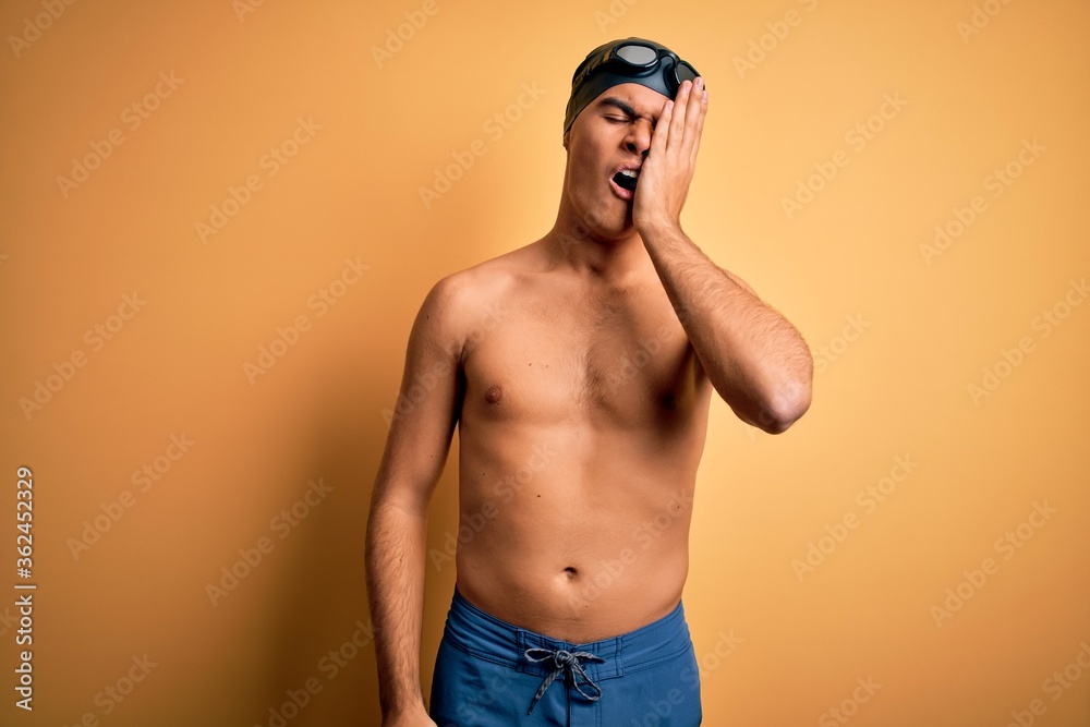 Young handsome man shirtless wearing swimsuit and swim cap over isolated yellow background Yawning tired covering half face, eye and mouth with hand. Face hurts in pain.