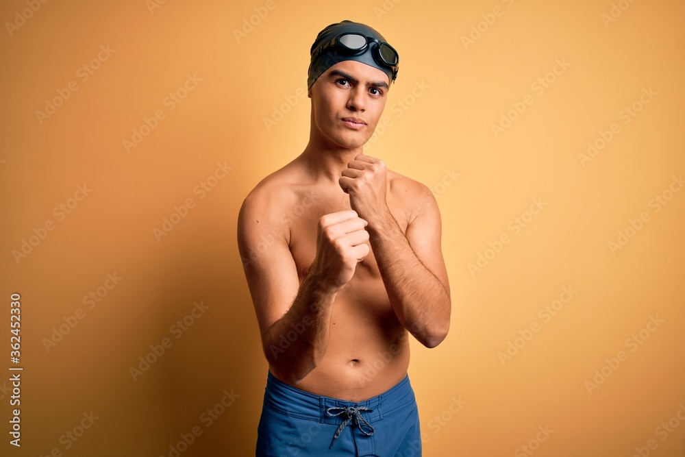 Young handsome man shirtless wearing swimsuit and swim cap over isolated yellow background Ready to fight with fist defense gesture, angry and upset face, afraid of problem