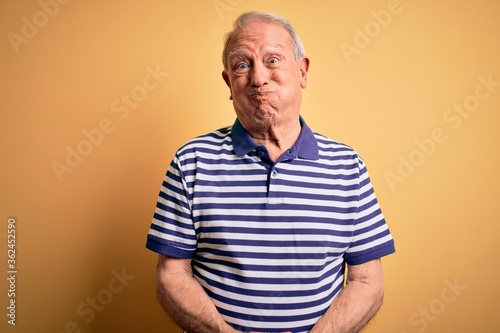 Grey haired senior man wearing casual navy striped t-shirt standing over yellow background puffing cheeks with funny face. Mouth inflated with air, crazy expression. © Krakenimages.com