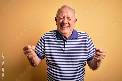 Grey haired senior man wearing casual navy striped t-shirt standing over yellow background very happy and excited doing winner gesture with arms raised, smiling and screaming for success. Celebration.