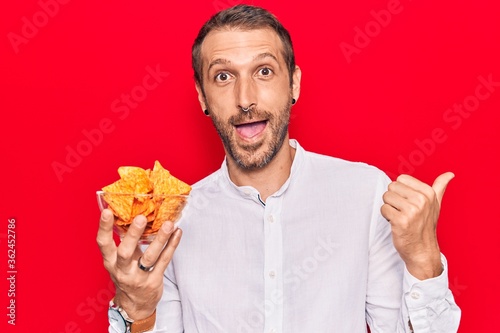 Young handsome man holding nachos potato chips pointing thumb up to the side smiling happy with open mouth