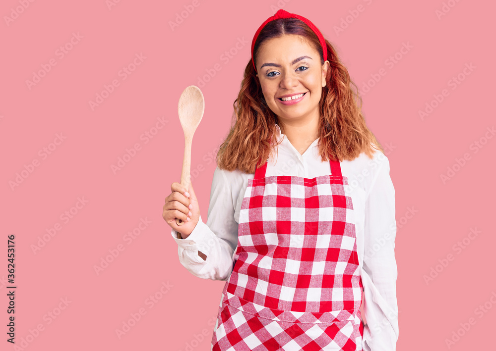 Young latin woman wearing apron holding wooden spoon looking positive and happy standing and smiling with a confident smile showing teeth Young latin woman wearing apron holding wooden spoon looking positive and happy standing and smiling with a confident smile showing teeth