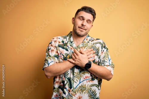 Young man with blue eyes on vacation wearing floral summer shirt over yellow background smiling with hands on chest with closed eyes and grateful gesture on face. Health concept.