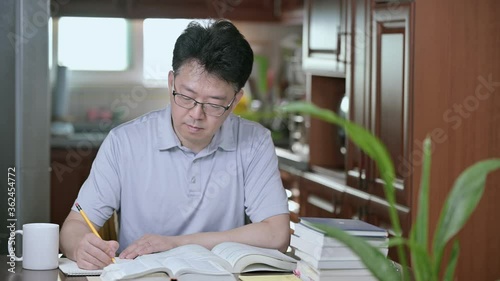 Asian middle-aged man sitting at desk at home, reading a book and studying.