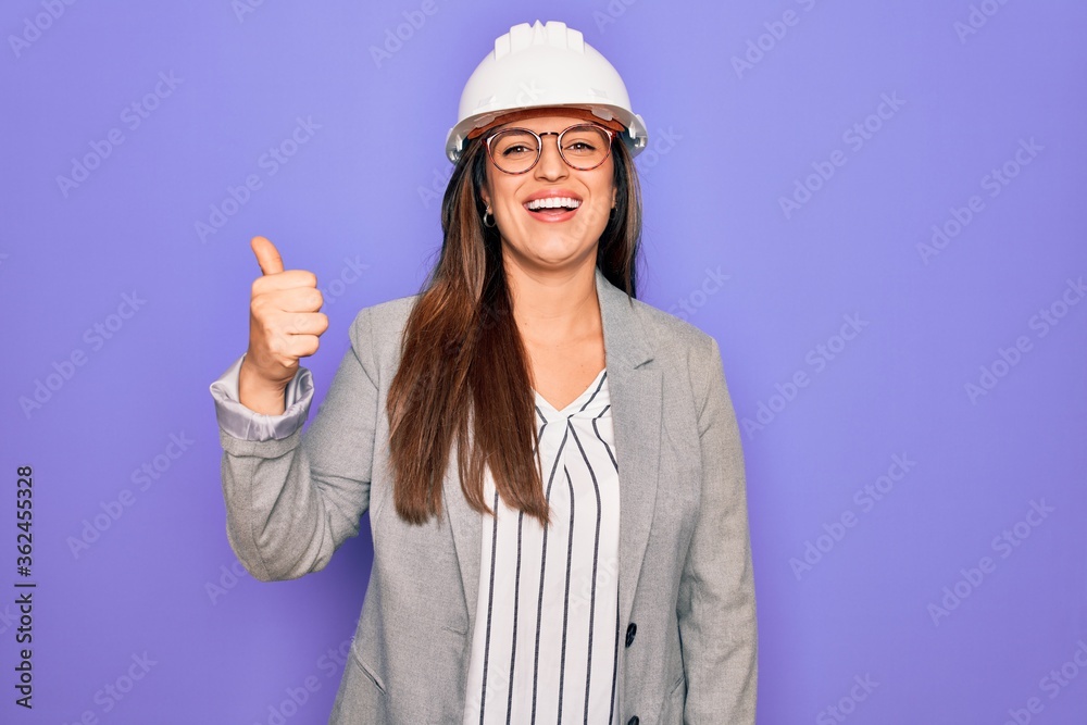 Professional woman engineer wearing industrial safety helmet over pruple background doing happy thumbs up gesture with hand. Approving expression looking at the camera showing success.