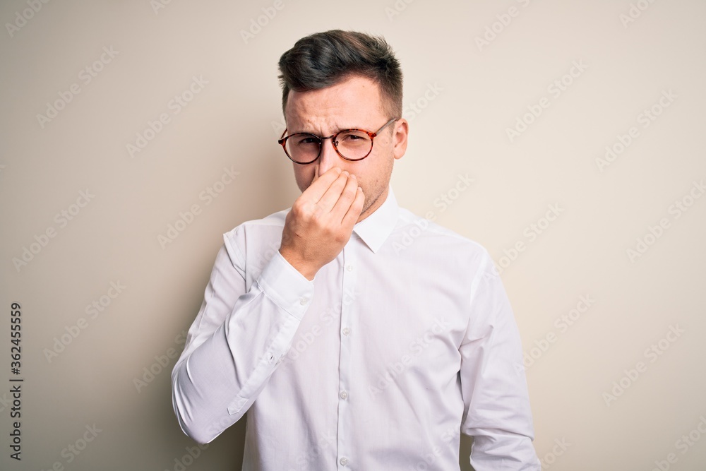 Young handsome business mas wearing glasses and elegant shirt over isolated background smelling something stinky and disgusting, intolerable smell, holding breath with fingers on nose. Bad smell