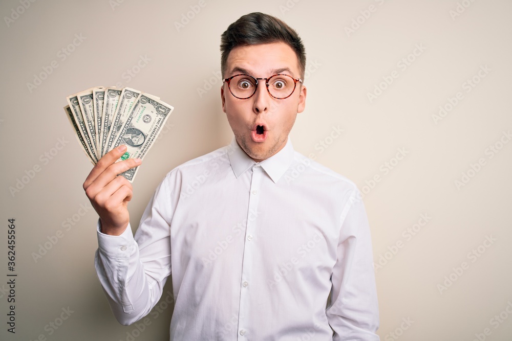 Young handsome caucasian business man holding bunch of one dollar banknotes scared in shock with a surprise face, afraid and excited with fear expression Young handsome caucasian business man holding bunch of one dollar banknotes scared in shock with a surprise face, afraid and excited with fear expression