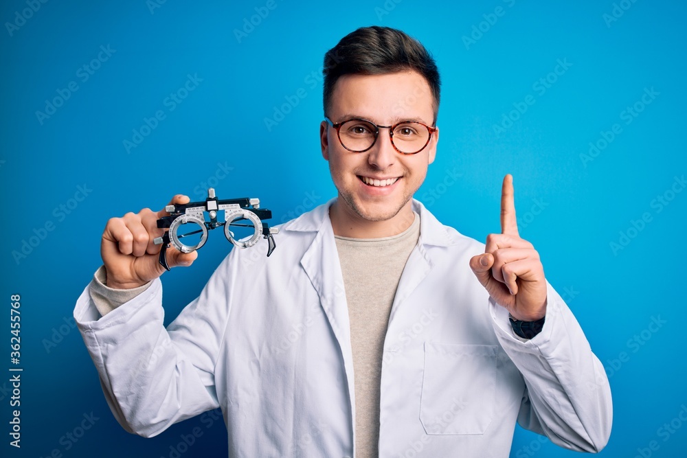 Young handsome professional optic man holding optometrical glasses over blue background surprised with an idea or question pointing finger with happy face, number one Young handsome professional optic man holding optometrical glasses over blue background surprised with an idea or question pointing finger with happy face, number one