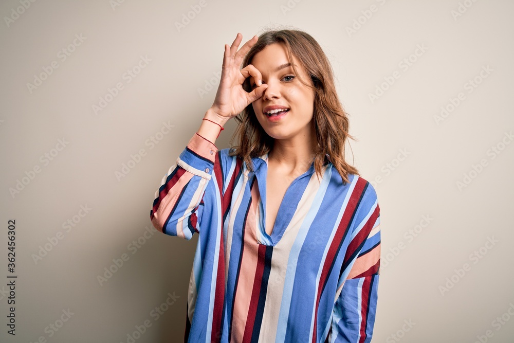 Young blonde girl wearing colorful striped shirt over isolated background doing ok gesture with hand smiling, eye looking through fingers with happy face. Young blonde girl wearing colorful striped shirt over isolated background doing ok gesture with hand smiling, eye looking through fingers with happy face.