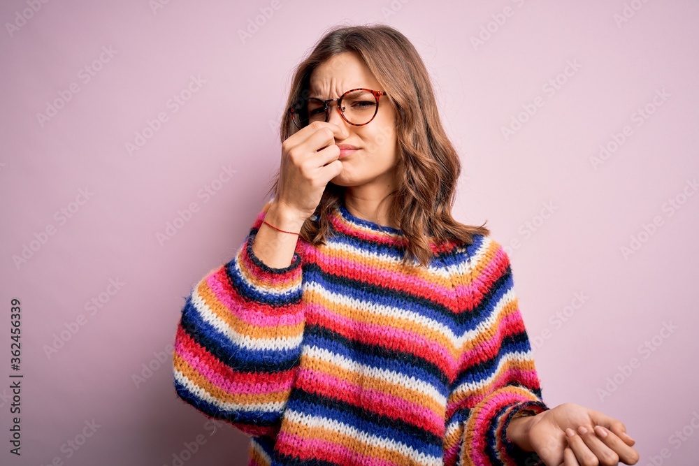 Young beautiful blonde girl wearing glasses and casual sweater over pink isolated background smelling something stinky and disgusting, intolerable smell, holding breath with fingers on nose. Bad smell Young beautiful blonde girl wearing glasses and casual sweater over pink isolated background smelling something stinky and disgusting, intolerable smell, holding breath with fingers on nose. Bad smell