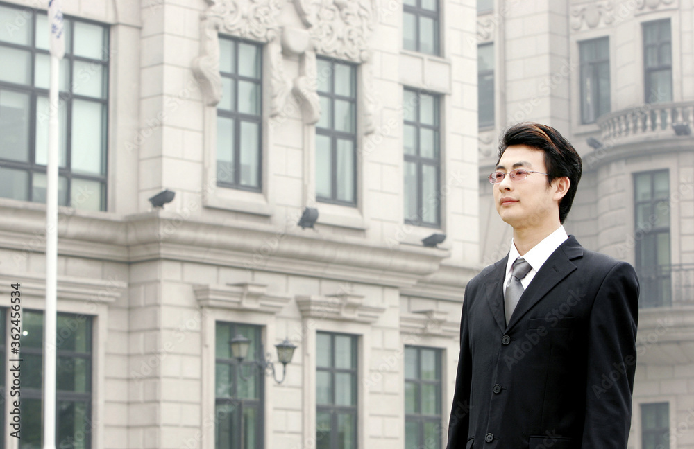 A bespectacled man in business suit standing in front of a building A bespectacled man in business suit standing in front of a building