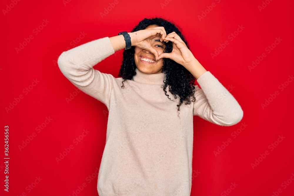 Young african american curly woman wearing casual turtleneck sweater over red background Doing heart shape with hand and fingers smiling looking through sign Young african american curly woman wearing casual turtleneck sweater over red background Doing heart shape with hand and fingers smiling looking through sign